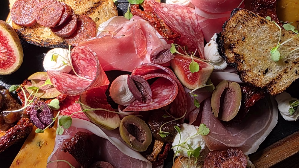 A colourful sharing board filled with cured meats, figs, olives, sun-dried tomatoes, whipped cheese and toasted sourdough, served under natural light at The Windmill.